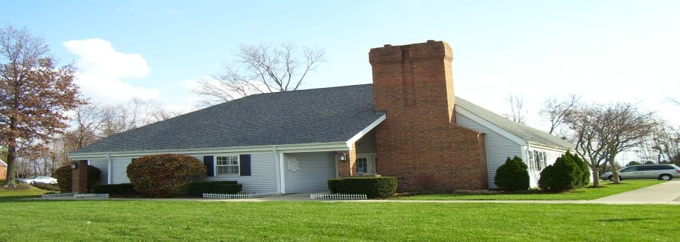 Low building with light siding and a large brick chimney behind a mown green lawn