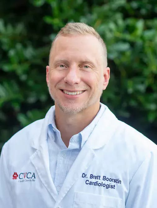 Smiling person in a white coat with a CVCA logo in front of green foliage