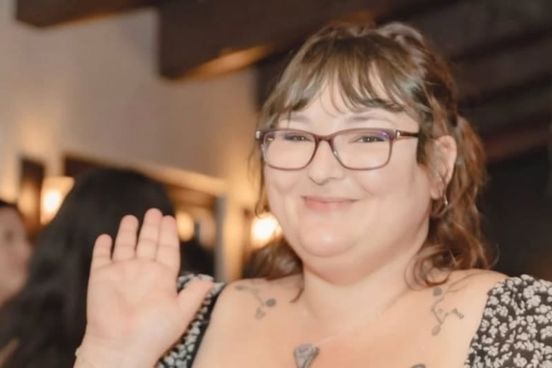 Smiling person with glasses and bangs waving at the camera in a dimly lit room