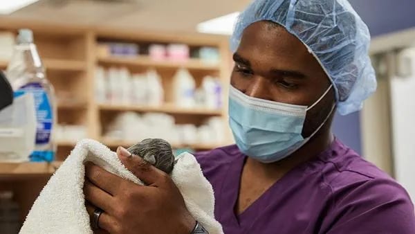 Person in purple scrubs, a blue hair cap and a face mask holding a small animal wrapped in a white towel
