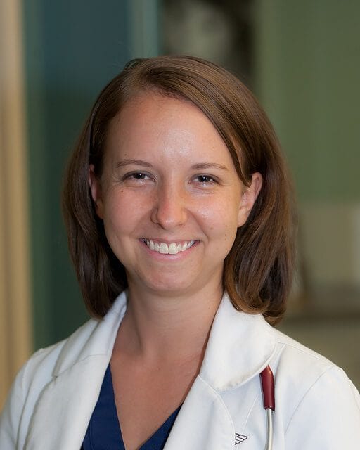 Smiling person with shoulder-length brown hair in a white coat over navy scrubs