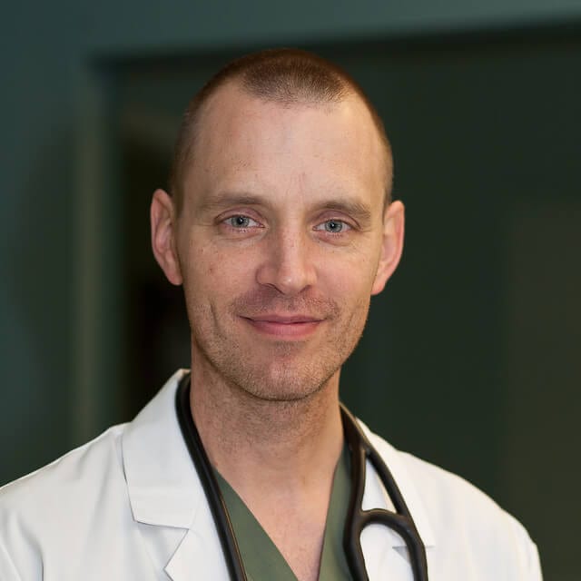 Person with short hair and a stethoscope around their neck in a white coat over green scrubs