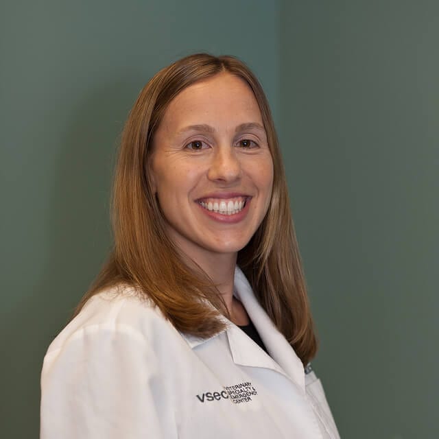Smiling person with long auburn hair in a white coat with a VSEC logo against a green-grey wall