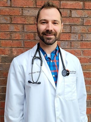 Smiling bearded person in a white coat and stethoscope in front of a brick wall