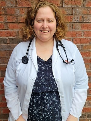 Smiling person with curly hair in a white coat and stethoscope by a brick wall