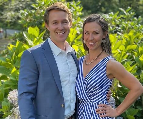 Smiling man in a blue blazer and woman in a striped dress standing in front of green foliage