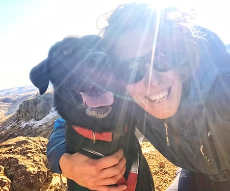 Smiling person in sunglasses hugging a black dog on a rocky hilltop in bright sun