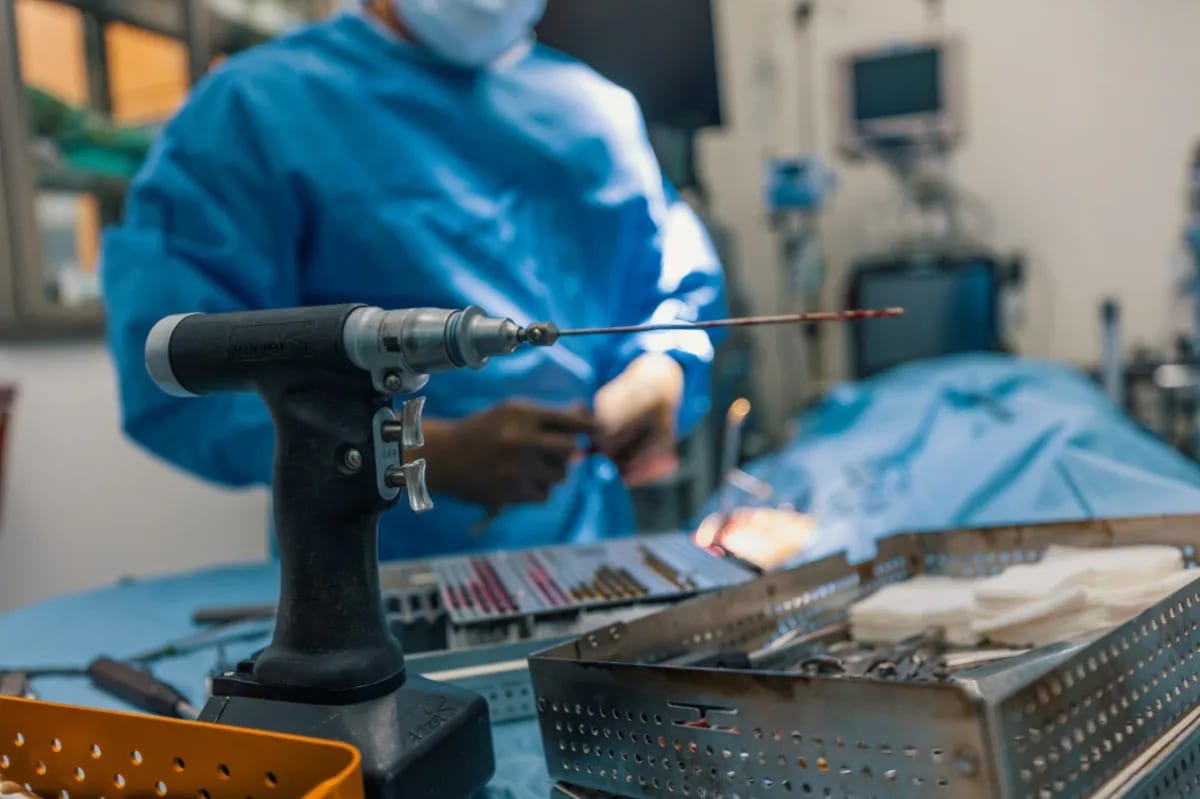 Person in blue gown and mask working at an operating table with a handheld power tool in front