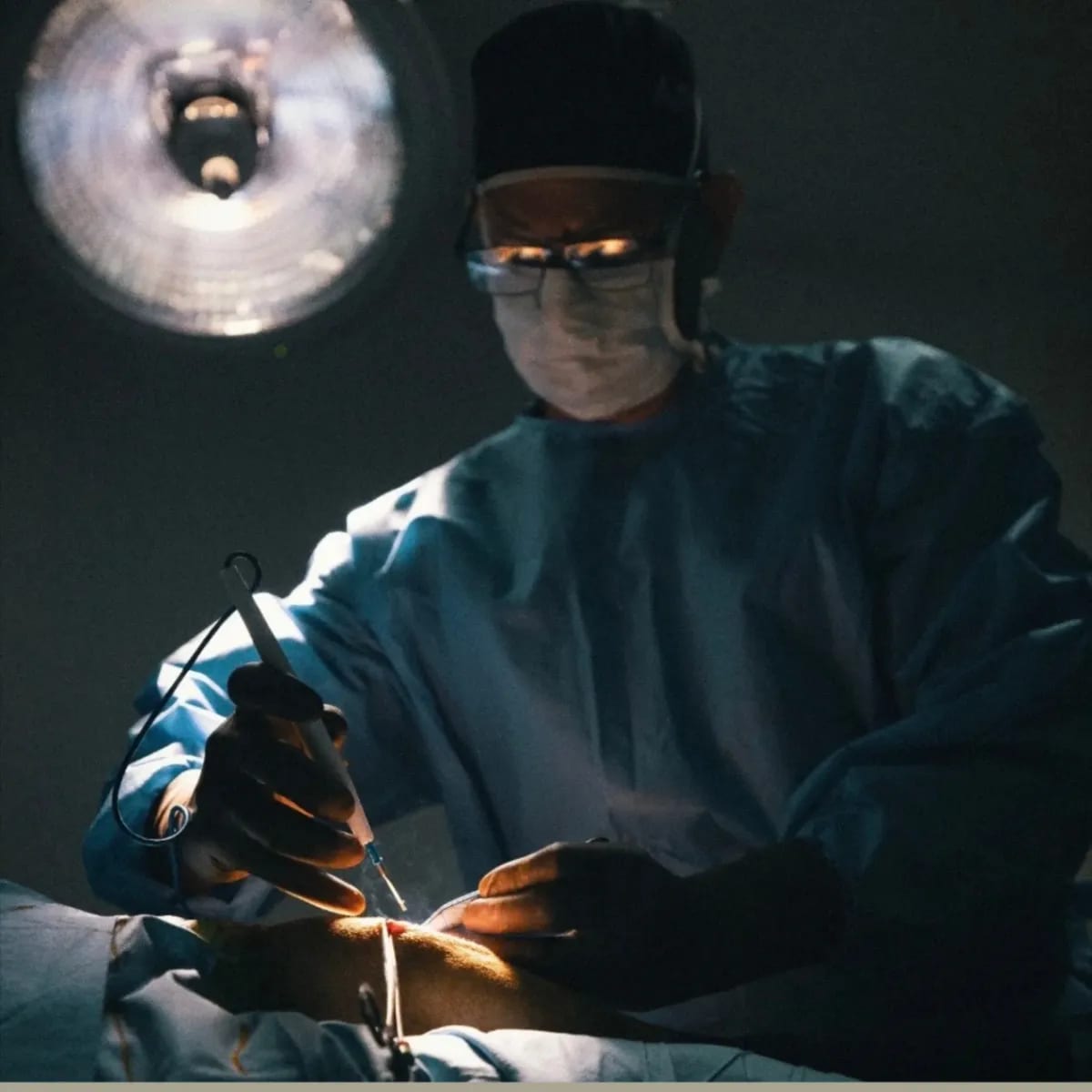 Person in a surgical cap, mask and gown holding forceps under a bright overhead lamp