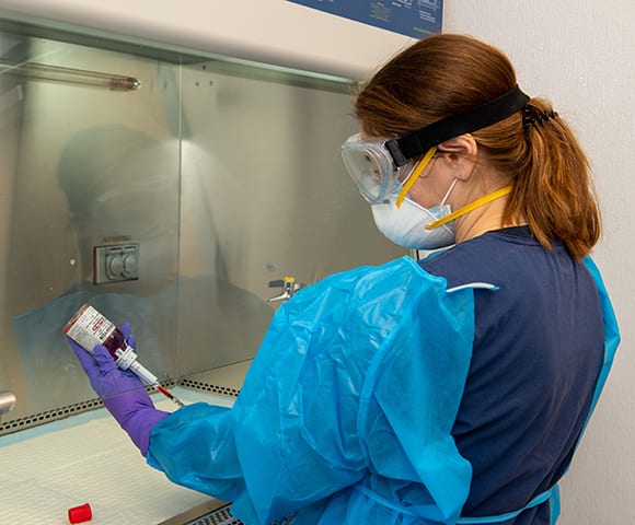 Person in a blue gown, goggles and mask holding a vial and syringe inside a steel cabinet
