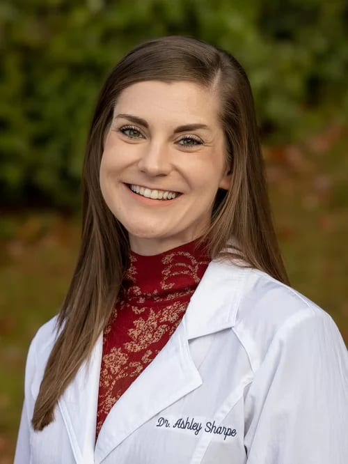 Smiling woman with long brown hair in a white coat over a red patterned top, outdoors