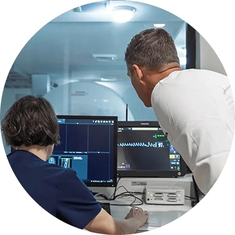 Two people at computer monitors in front of a large ring-shaped machine behind a glass window