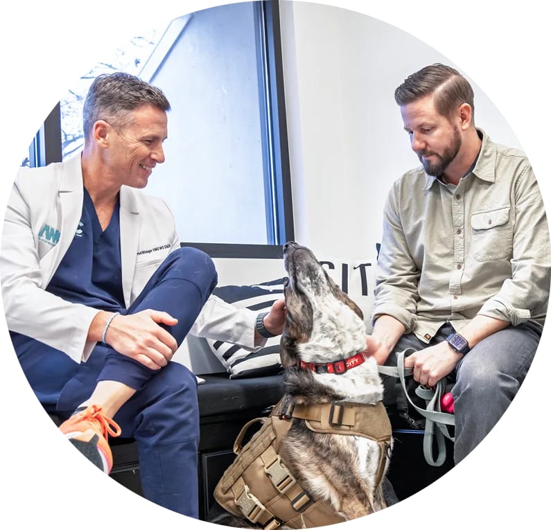 Two men seated beside a dog in a tan harness, one in a white coat over navy scrubs