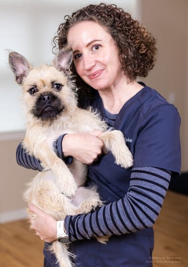 Smiling woman with curly hair in a navy top holding a small tan dog with a dark face
