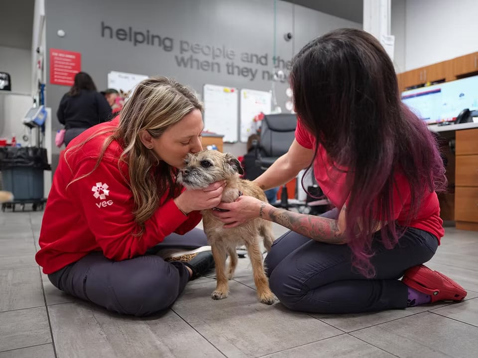 Two people in red tops kneeling on a floor with a small tan dog, one kissing its head