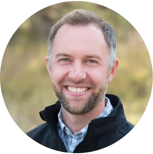 Smiling man with a short beard wearing a plaid collared shirt and black jacket outdoors