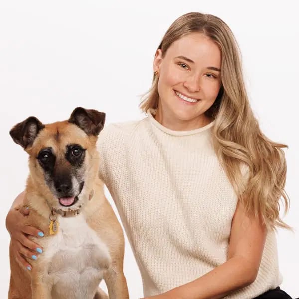 Smiling woman with long blond hair in a cream knit top with her arm around a tan dog with a dark muzzle