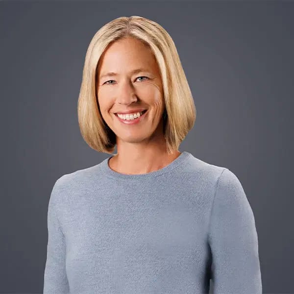 Smiling woman with chin-length blond hair in a light blue sweater against a dark grey background