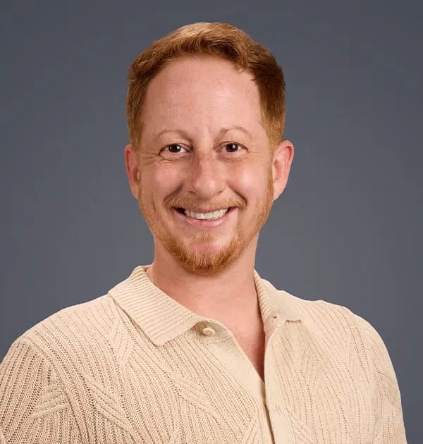 Smiling man with short red hair and a goatee in a cream knit polo shirt against a grey background