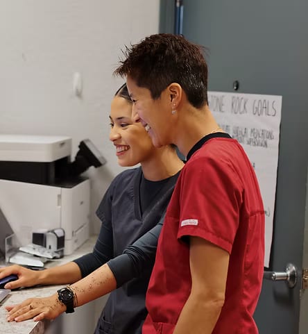 Two people in red and grey scrubs smiling as they look down at a counter