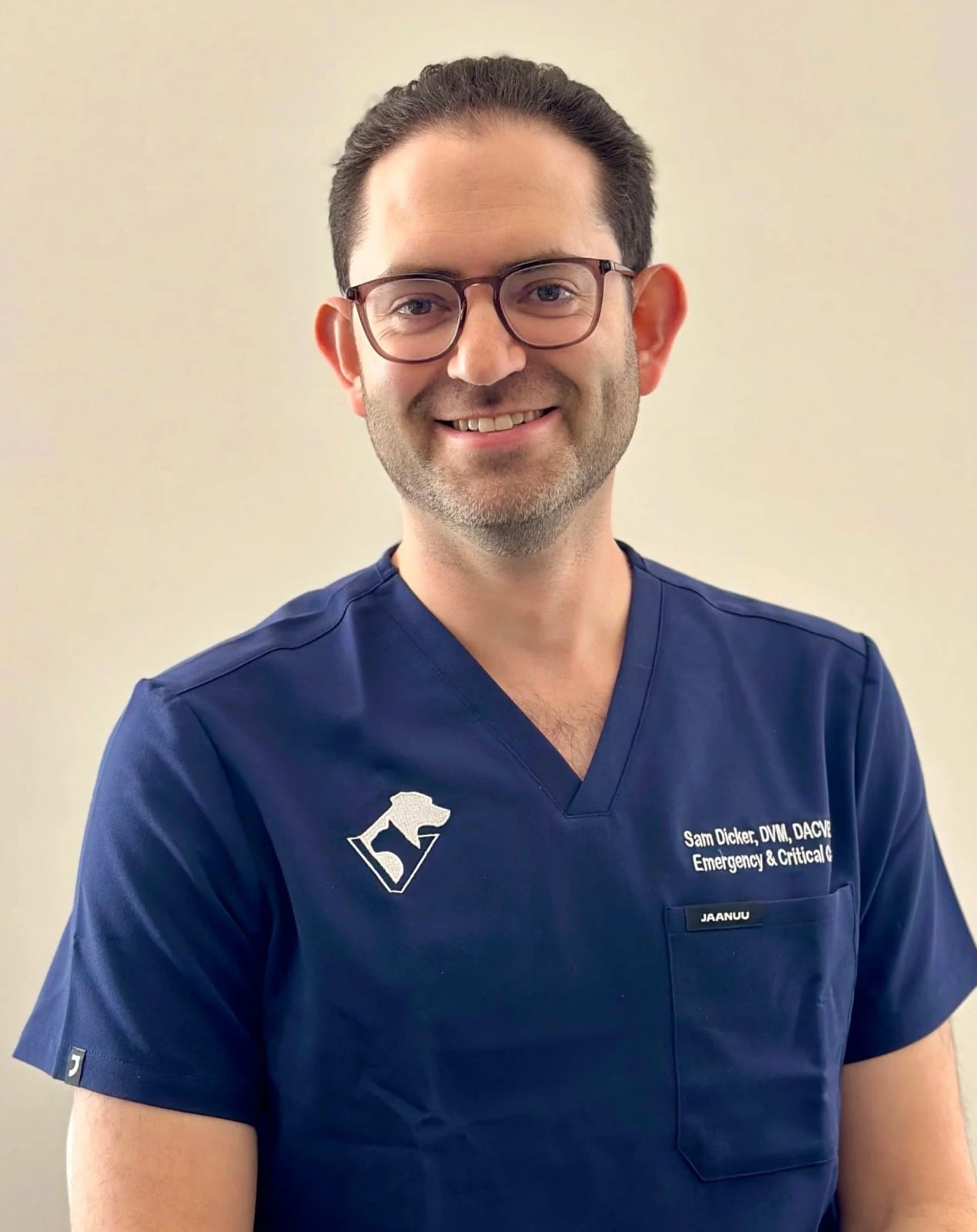 Smiling bearded person with glasses in navy scrubs with a dog logo, against a plain cream wall