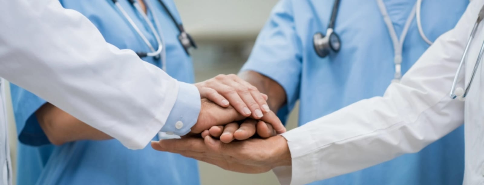 Hands stacked together in the centre of a circle of people in teal scrubs and white coats wearing stethoscopes
