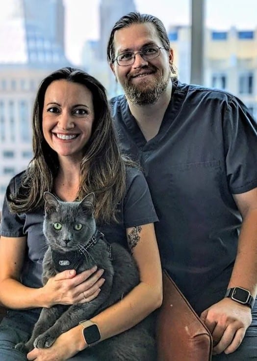 Two smiling people in grey scrubs with a grey cat, in front of a window with city buildings