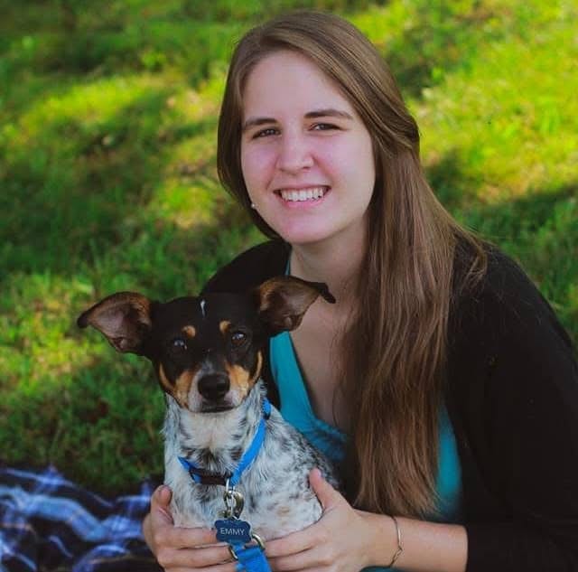 Smiling person with long brown hair holding a small dog with a blue collar on a lawn