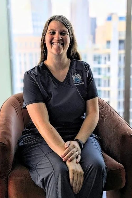 Smiling person in dark grey scrubs sitting in a rust-coloured armchair in front of a window with city buildings