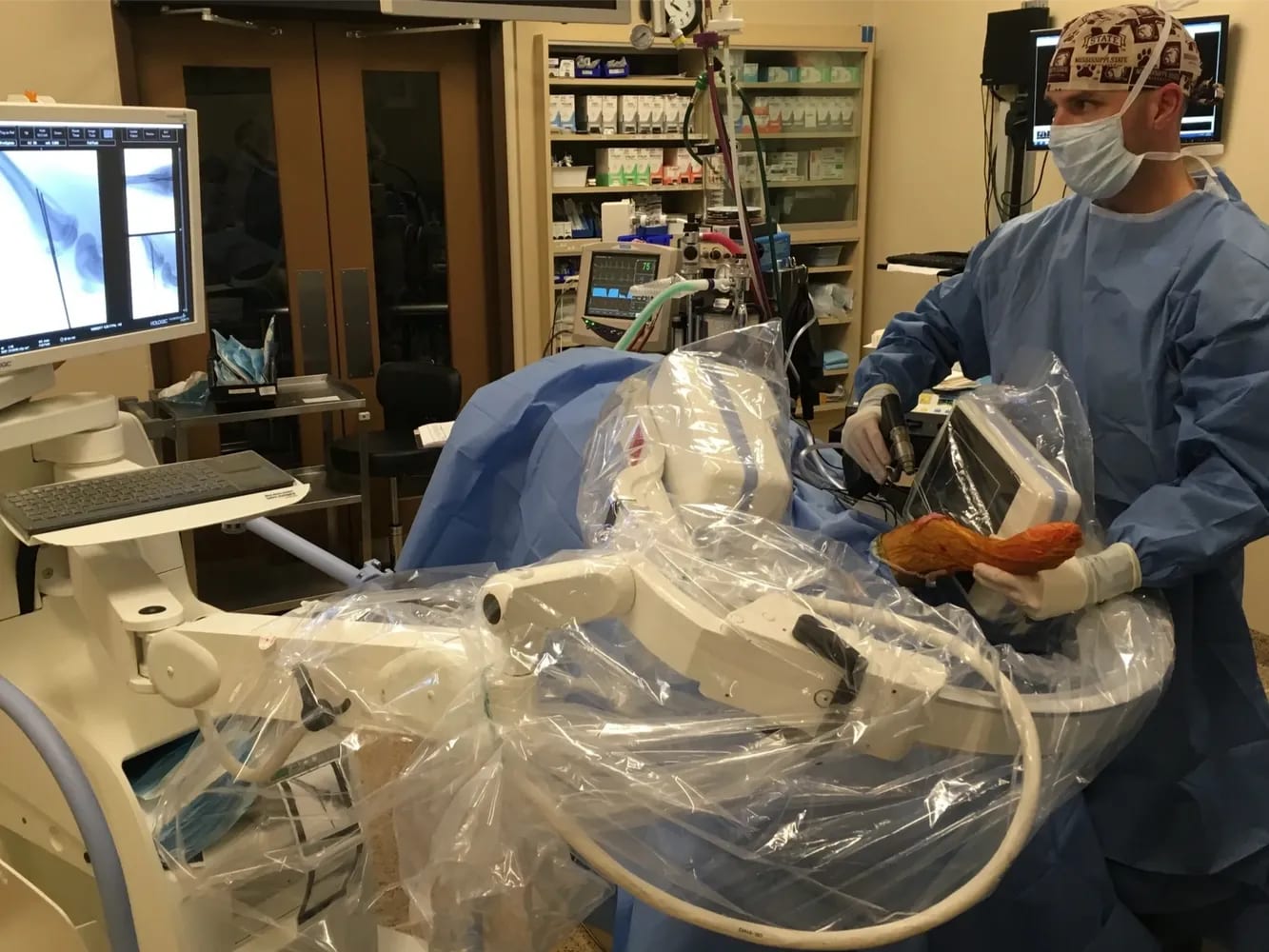 Person in a blue gown, cap and mask beside a large plastic-draped imaging machine in a clinical room