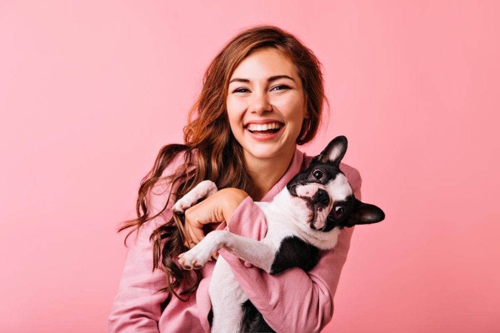 Smiling woman in a pink top holding a black and white dog against a pink background