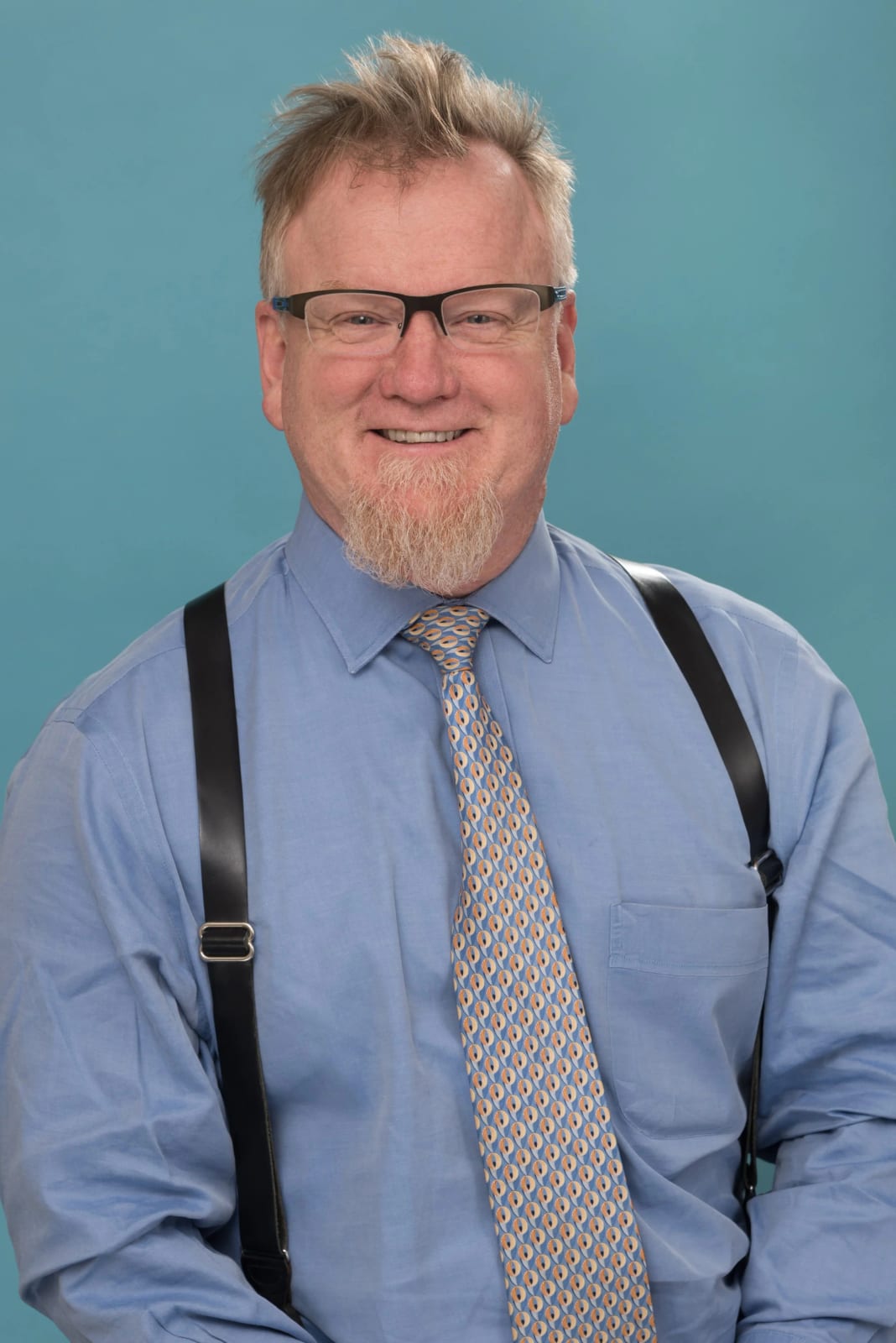 Smiling man with glasses, a light goatee, a blue shirt, patterned tie and black suspenders
