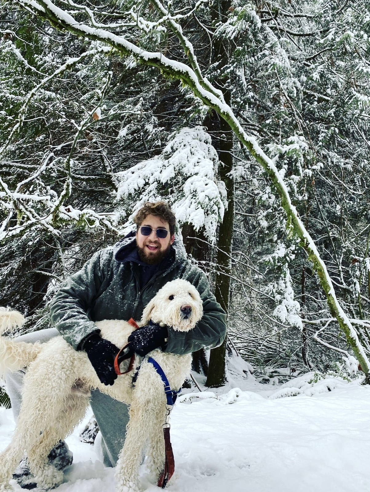 Bearded man in sunglasses holding a large fluffy white dog in a snowy forest