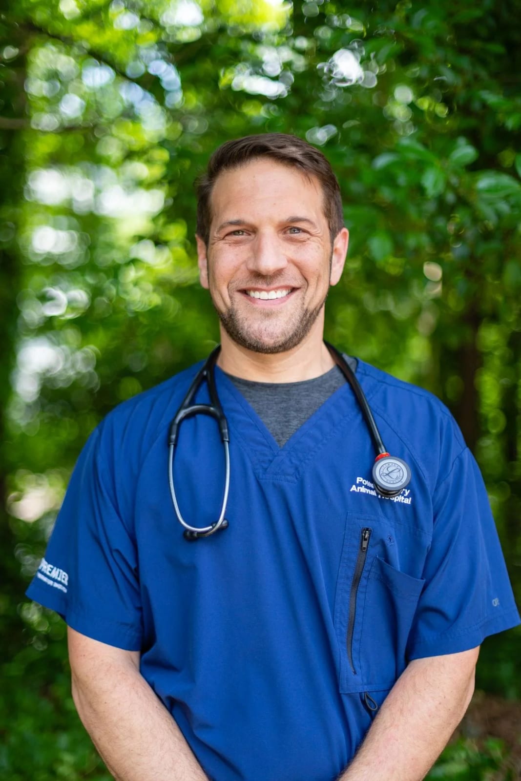 Smiling man with stubble in blue scrubs and a stethoscope standing in front of green trees