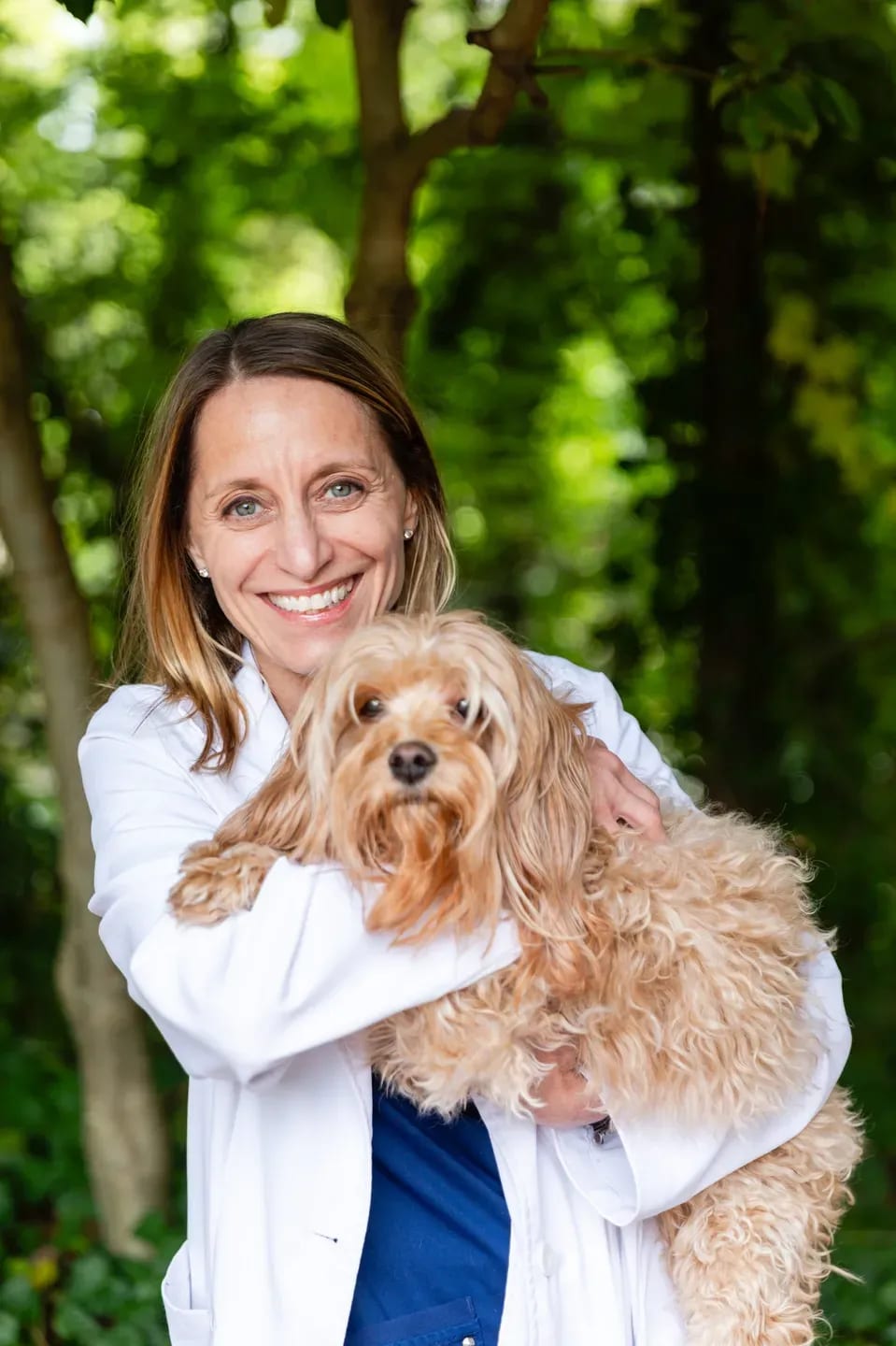 Smiling woman in a white coat holding a fluffy apricot dog in front of green trees