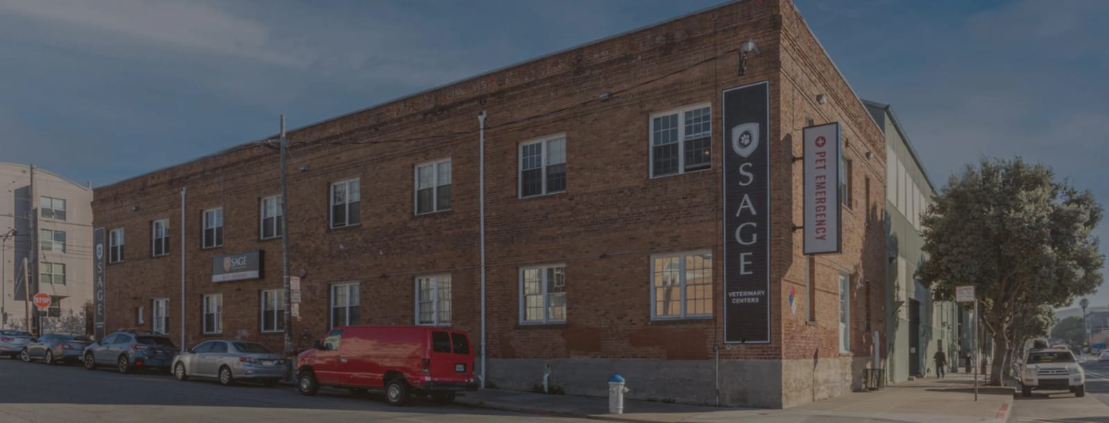 Long red brick building with SAGE and Pet Emergency signs, a red van and parked cars on the street