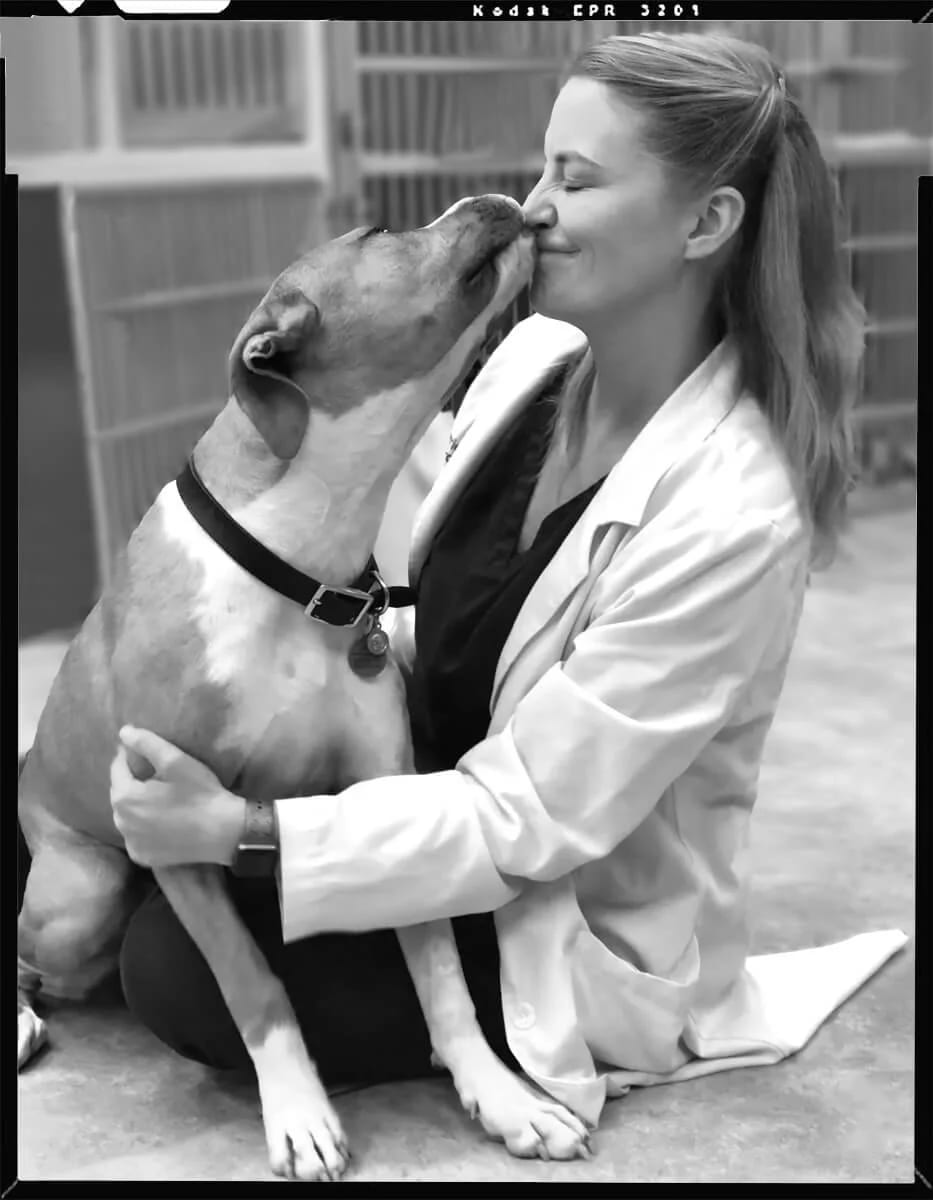 Black and white photo of a large dog licking the face of a kneeling woman in a white coat