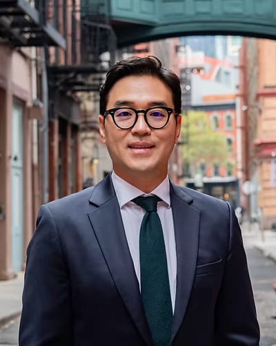 Man with round glasses, a navy suit and a green tie standing on a city street