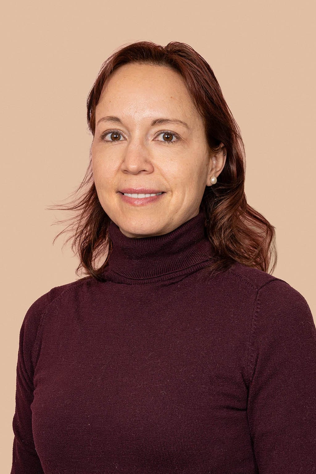 Smiling woman with shoulder-length auburn hair in a maroon turtleneck on a peach background