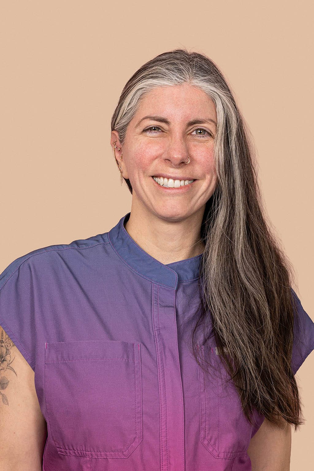 Smiling person with long grey-streaked hair in a purple and pink scrub top on a peach background