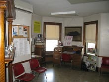 Small low-resolution room with red chairs, a wooden desk, a bulletin board and two windows