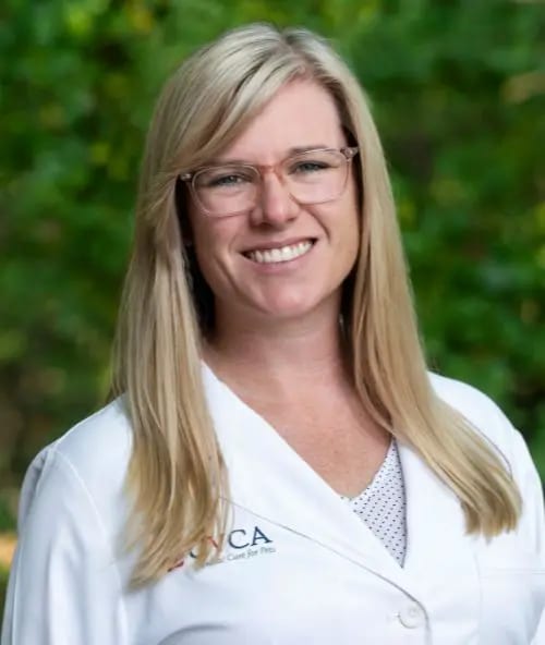 Smiling woman with long blonde hair and pink glasses in a white coat with a CVCA logo in front of trees