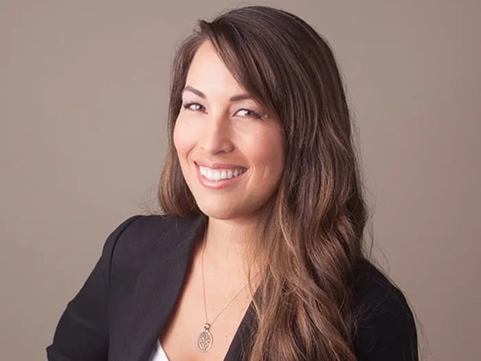 Smiling woman with long brown hair wearing a black blazer and pendant necklace against a taupe background