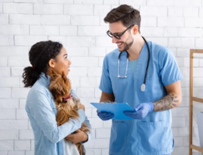 Smiling person with glasses in light blue scrubs holding a clipboard beside a woman holding a small dog
