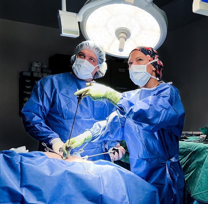 Two people in blue gowns, masks and caps holding long instruments under a surgical light