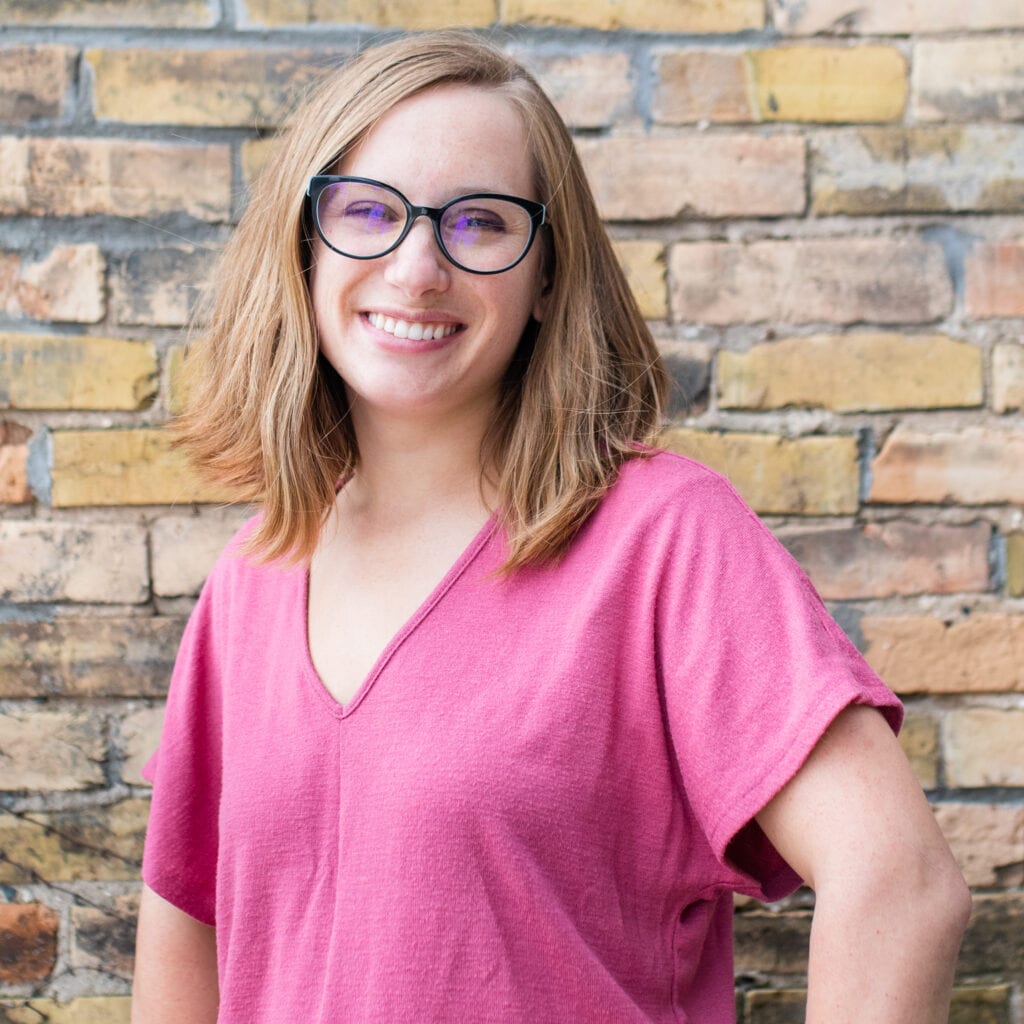 Smiling person with glasses and shoulder-length hair in a pink top against a yellow brick wall