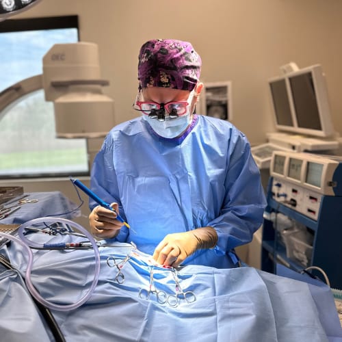 Person in a patterned scrub cap, mask and blue gown working at a draped table with metal instruments