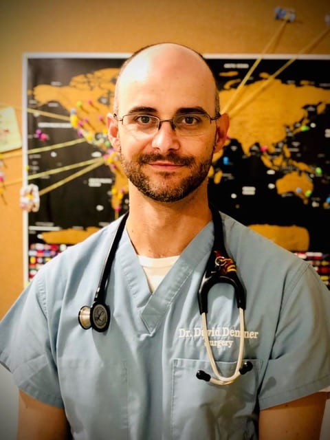 Bald person with glasses and a beard in grey-blue scrubs and a stethoscope in front of a world map