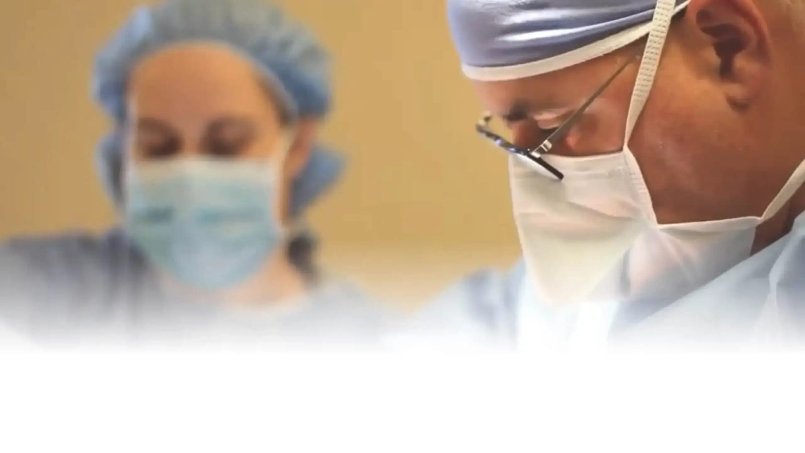 Two people in surgical caps and face masks, one wearing glasses, with a white fade at the bottom