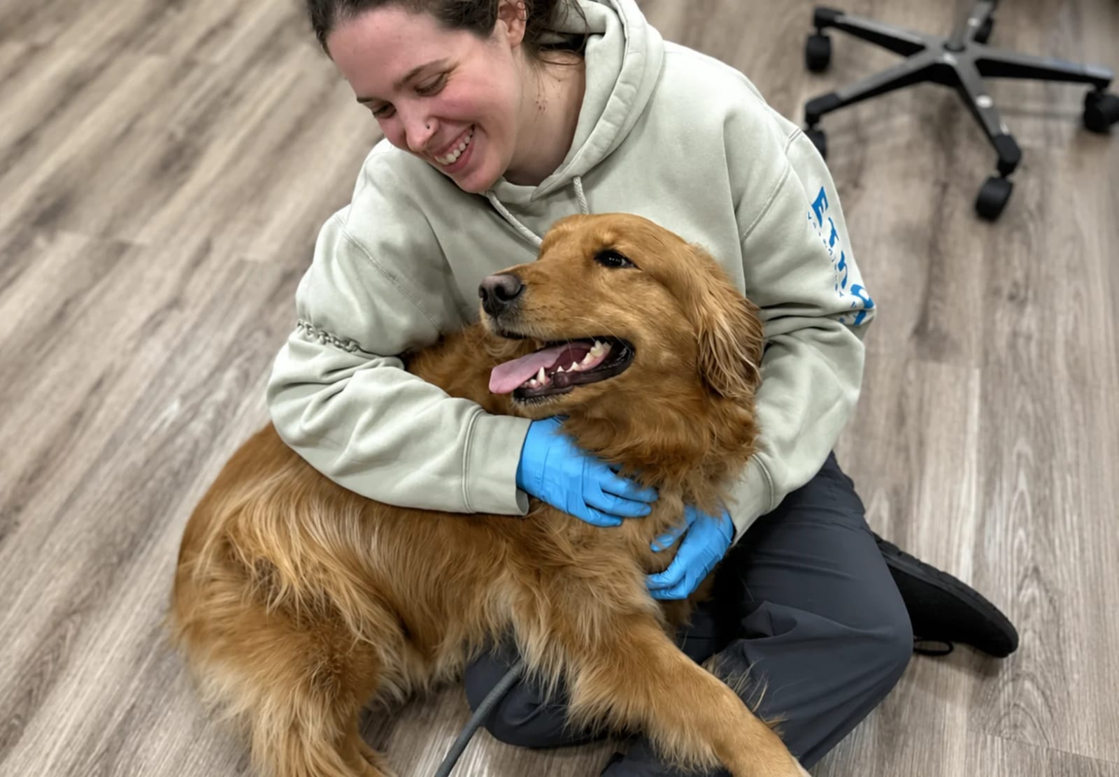 Smiling person in a green hooded sweatshirt and blue gloves hugging a golden dog on a wood floor